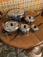 All six stainless steel cooking pots with lids arranged on wooden table, showing range of sizes and styles
