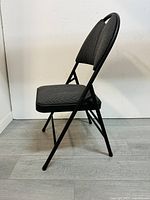 Side profile view of a black foldable chair with cushioned seat and backrest against a white wall and light wooden floor.
