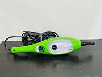 Green handheld H2O DualBlast steam cleaner head with attached power cord lying flat on black surface against white wall.