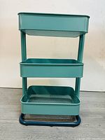 Front view of the 3-tier teal metal kitchen utility cart, showing three storage levels with two solid shelves and one mesh-bottom shelf, supported on caster wheels with a black rounded metal base.
