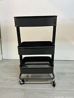 Front view of black metal kitchen rolling utility cart with three tiers and caster wheels on a white background.