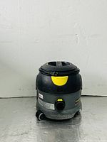 Front-right side view of the Karcher Professional T12/1 vacuum cleaner showing the cylindrical black and gray body with yellow details, sitting on wheels on a metal surface.