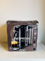 Front view of the original box showing image of Nespresso VertuoPlus Deluxe coffee machine by DeLonghi and coffee cup, box is worn and creased.