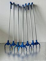 Photo showing seven Oates brand 1-meter nippers reachers standing upright in a row with blue claw gripping heads and blue handles against a white wall on a metallic surface.