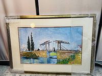 Front view of framed wall art painting showing a stone bridge over a body of water with blue sky and two tall trees on the left side. The piece is covered in protective bubble wrap with the frame visible.