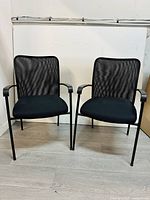 Front view of two black mesh office chairs with cushioned fabric seats, metal legs and armrests, shown on light colored floor against a white wall.