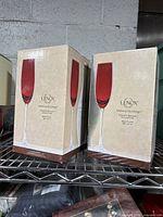 Two unopened beige boxes each containing 4 Lenox red flute glasses from the Tuscany Holiday Collection on a metal shelf.