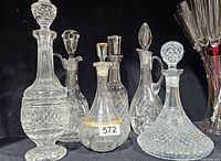 Photo of 6 crystal cut glass decanters showing variety of shapes and stoppers on black background.