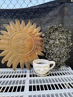 Full view of the lot showing the metal sun sculpture, Green Man resin wall plaque, and ceramic face mug planter with saucer on a white grid shelf against a black mesh backdrop.