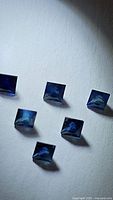 Six deep blue to teal green princess cut sapphires on a white background showing size and color variation