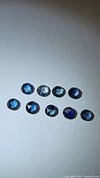 Photo showing 9 loose blue sapphires with rose cut and varying blue hues arranged in two rows on white background.