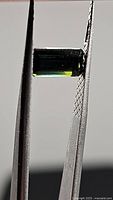 Close-up photo of the bright green octagon cut tourmaline held by tweezers showing its color and facets.