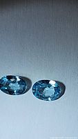 Two oval cut, deep seafoam blue Cambodian zircon gemstones placed on a white surface, showing clarity and color.