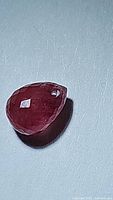Close-up photo of a pinkish red briolette cut ruby gemstone with a drilled hole near the top for jewelry attachment, showing faceted surface and transparency.