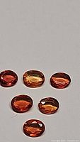 Six oval faceted natural fire orange red sapphires arranged on a light background showing color and cut details.
