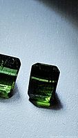 Close-up image of two natural green tourmaline gemstones with a rectangular fancy cut, showing facets and natural characteristics.
