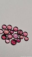 Photo showing 18 natural round cabochon sapphires in various pink and red raspberry shades, sizes approximately 2mm to 2.5mm, arranged on a light background.