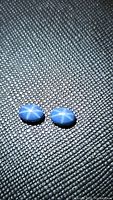 Pair of oval cabochon natural star sapphires showing a distinct 6-ray star effect on textured dark background.