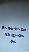 Eight pear cut, purple-blue tanzanite gemstones arranged on a flat neutral background under lighting to show facets and color