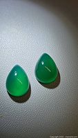 Two vivid deep green pear-shaped onyx cabochons on a white textured surface, showing smooth domed finishes and translucency.