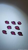 Top view of eight oval cut deep fuchsia pink purple rhodolite garnet gemstones arranged on white background with spotlight.