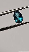 Oval shaped natural topaz gemstone held with tweezers showing deep aqua blue-green coloration and facets