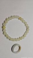 Photo shows the natural white jade stretchy bracelet made of round beads arranged in a circle and a solid polished white jade ring with yellowish markings beside it.
