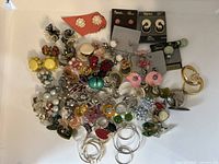 Top-down view of numerous pairs of earrings assorted on a white surface including metal hoops, colorful resin and bead floral, geometric and abstract designs with some pairs on original branded cards.