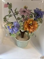 Full view of multicolor stone flower arrangement with multiple flower types and green leaves in light green ceramic pot with textured stone base.