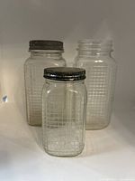 Front view of three waffle-patterned glass jars: two large and one small, two with metal lids and one lidless.