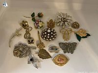 Collection of 15 assorted vintage brooches laid out on a beige surface, showing various floral, insect, and decorative designs in gold, silver, pearl, and enamel tones.