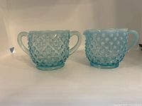 Two light blue opalescent hobnail glass pieces side by side, viewed from front - creamer with pouring spout, sugar bowl with two handles