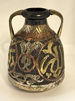Front-side view of the vintage metal jug showing repousse Egyptian motifs with gold, copper, and silver tones, two handles, black neck, and minor surface wear.