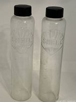Full view of both clear cylindrical glass bottles with black screw caps, showing embossed Shell Oil Company logos.