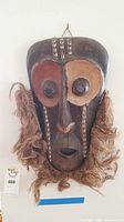Full view of the wooden African mask showing facial carving, eye color patterns, fiber fringe, and shell decoration on forehead.