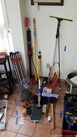Photo showing assortment of long handled brushes, a plunger, a mop, adjustable metal table legs, and various cleaning tools laid on tiled floor near white wall