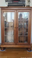 Front view of the wood cabinet with two glass doors, showing glassware inside. Keyhole lock and brass hinges visible. Cabinet stands on rounded feet.