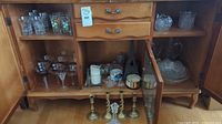 Wooden cabinet shelves displaying various glassware including glasses, goblets, candleholders, and a miniature Eiffel Tower decoration, as well as brass candleholders and pillar candles on bottom shelf.