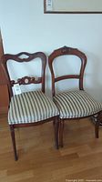 Two wooden frame chairs with carved backs and striped upholstered seats next to each other against a wall.