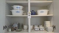 Full shelf view showing white CorningWare casserole dishes with blue floral Cornflower Blue pattern, white ceramic ramekins on right, and glass bowls and divided plates on left.
