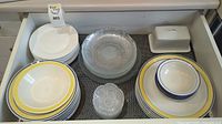 Top view of white dining plates with yellow and blue rims, clear glass salad plates, small glass bowls, and a ceramic butter dish inside a drawer.