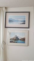 Two framed landscape artworks hanging on a beige wall; top print features blue misty seascape mountains, bottom original art shows detailed mountain lake scene.