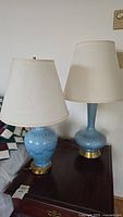 Two blue ceramic table lamps with white shades standing on a wooden surface near a quilt.