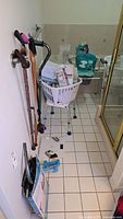 Photo of bathroom floor showing a white shower chair, three walking canes hanging on the wall, a white scale on floor, a set of blue hand weights, a laundry basket with toiletries, and a metal wall bar along the wall.
