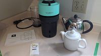 Photo of Nespresso Vertuo Plus coffee maker in turquoise and black with instruction manual, power cord, stainless steel teapot, and white ceramic sugar pot.