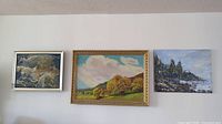 Three original paintings displayed on a wall: abstract sea scene, autumnal landscape with trees in golden frame, rocky coastal scene.