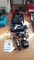 Photo showing a collection of power tools including Bosch drills, Porter Cable jig saw, orbit sander, soldering and heat tools, along with chargers and batteries placed on floor.