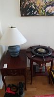 Photo showing rectangular wood table with ceramic lamp on top and damaged wooden ship's wheel barometer on adjacent round table