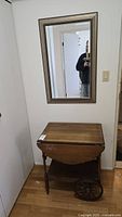 View of the tea cart and the mirror mounted above it. The tea cart has visible wood grain and the mirror reflects part of the room.