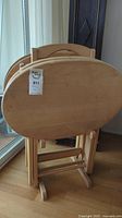 Set of 4 oval solid wood TV dinner tables stacked folded next to window.